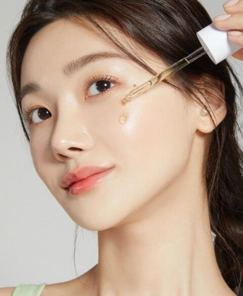 Woman applying facial serum with a dropper on her cheek, showcasing glowing and hydrated skin.